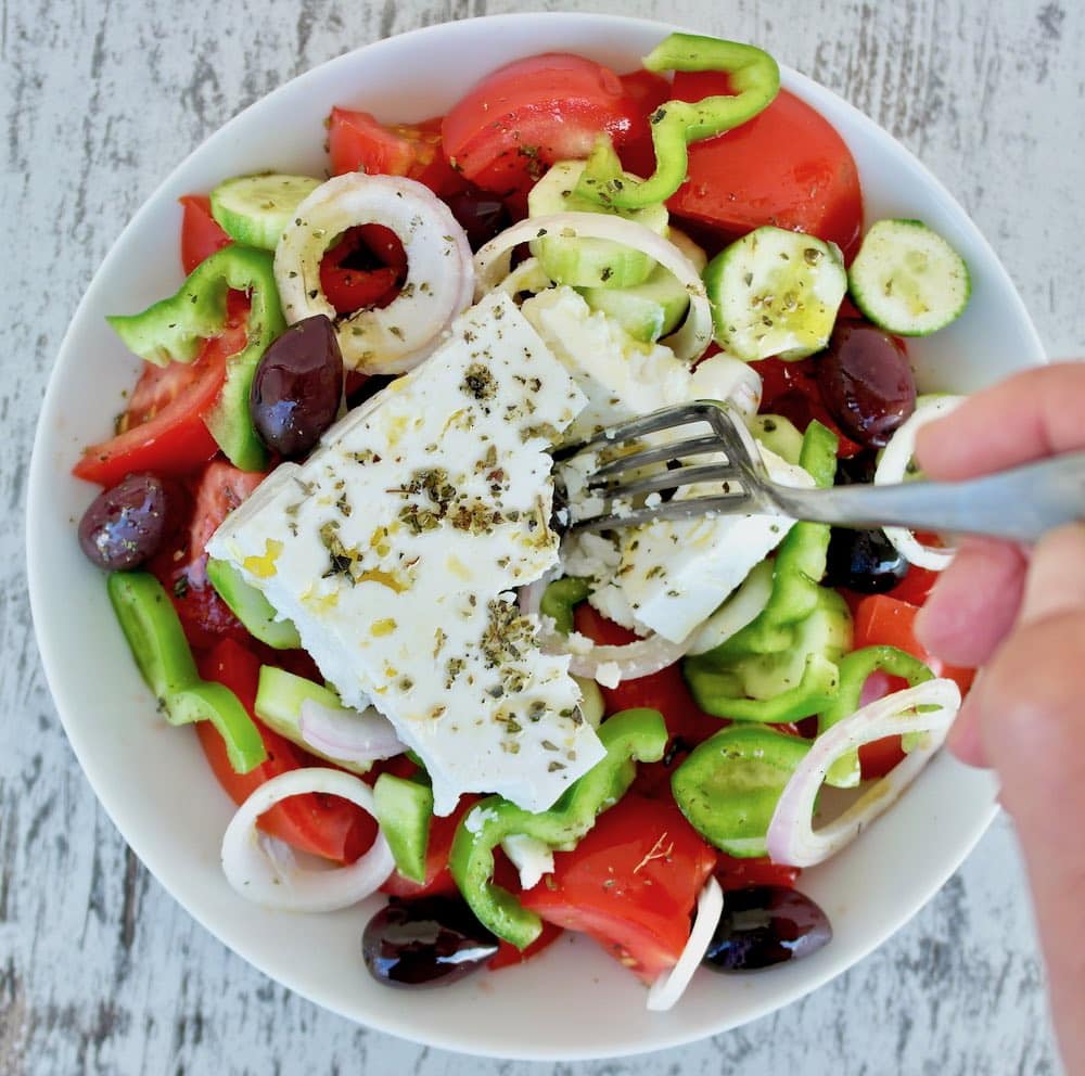 A traditional Greek salad made with tomatoes, cucumbers, olives, pepper, onion and feta in a white bowl.